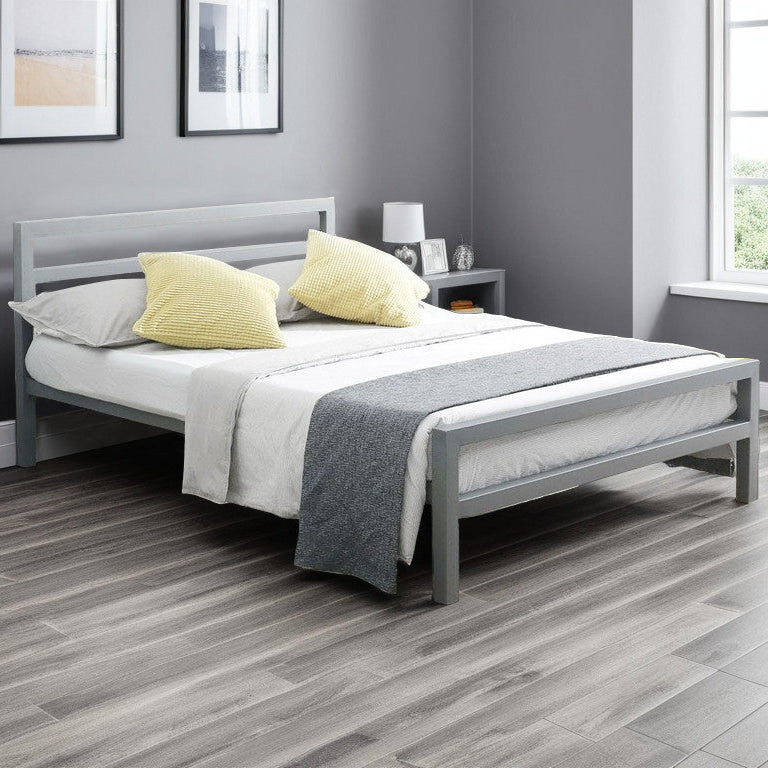 A silver metal bed frame with a simple, modern design, placed in a neatly arranged bedroom with pillows on the bed and a window in the background.