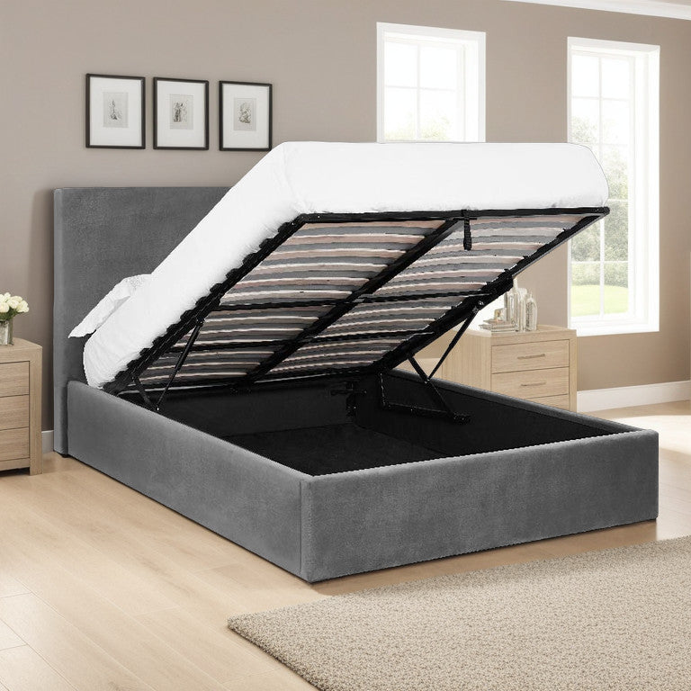 A gray velvet-upholstered Lisbon Fabric Ottoman Bed Frame with a hydraulic lift mechanism and a tall headboard, displayed in a stylized bedroom setting.