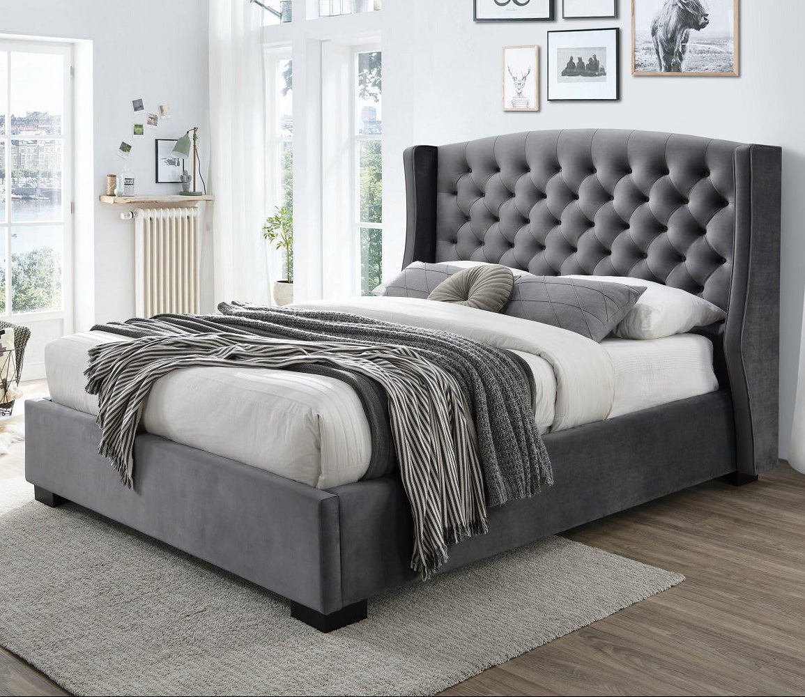 A grey upholstered bed frame with a tufted headboard and winged design, placed in a bedroom setting with bedding and a shawl added for a cozy look.
