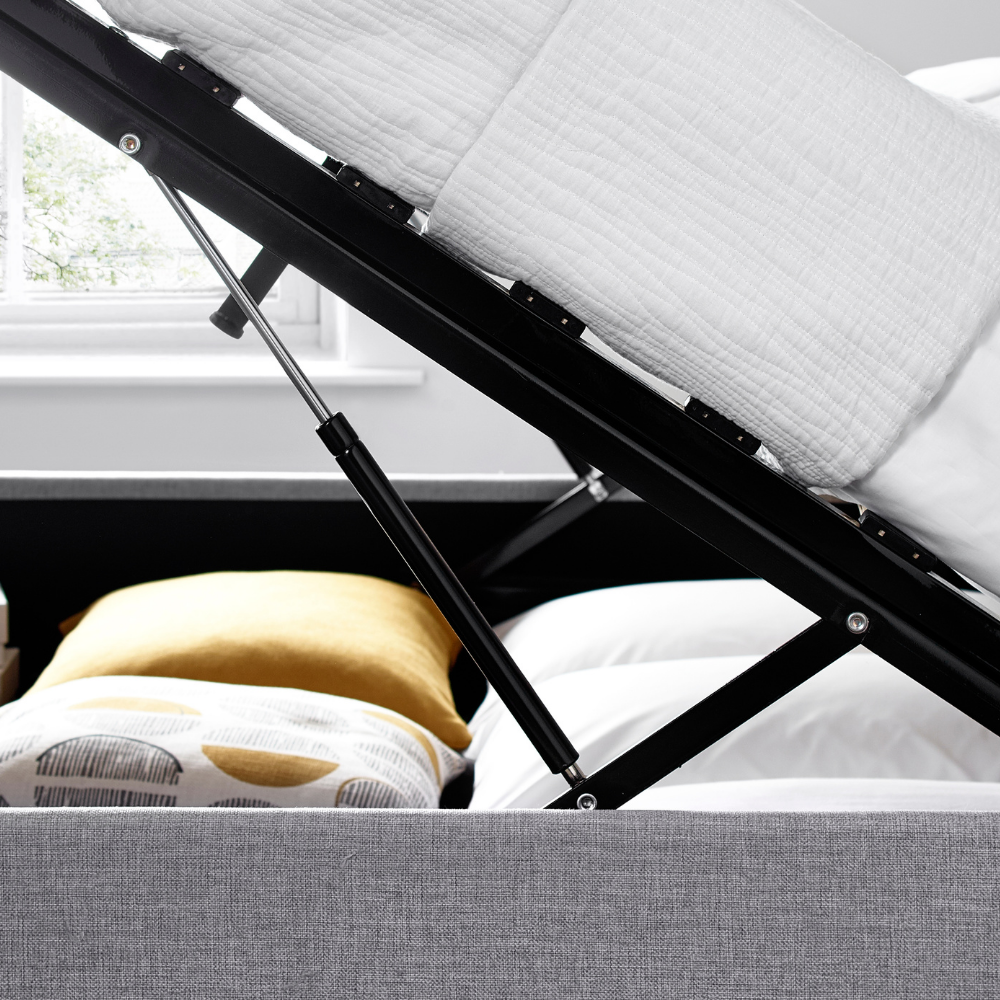 Grey ottoman bed with black metal frame in a room with a window and pillows.