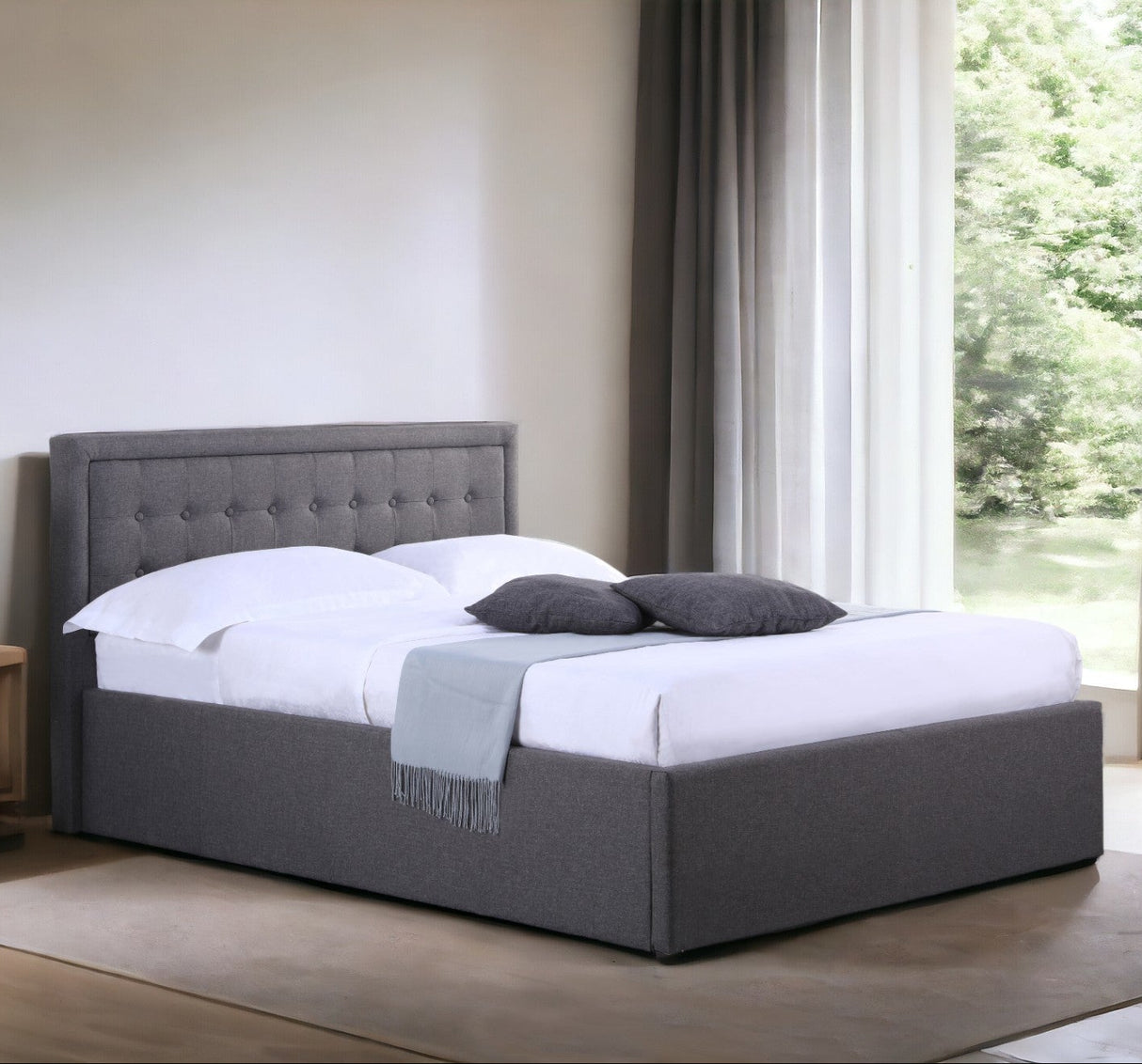 A double-sized Houston Fabric Ottoman Bed Frame in gray, featuring a buttoned headboard, with white bedding and two gray pillows, placed in a room with a large window and a side table with a lamp.