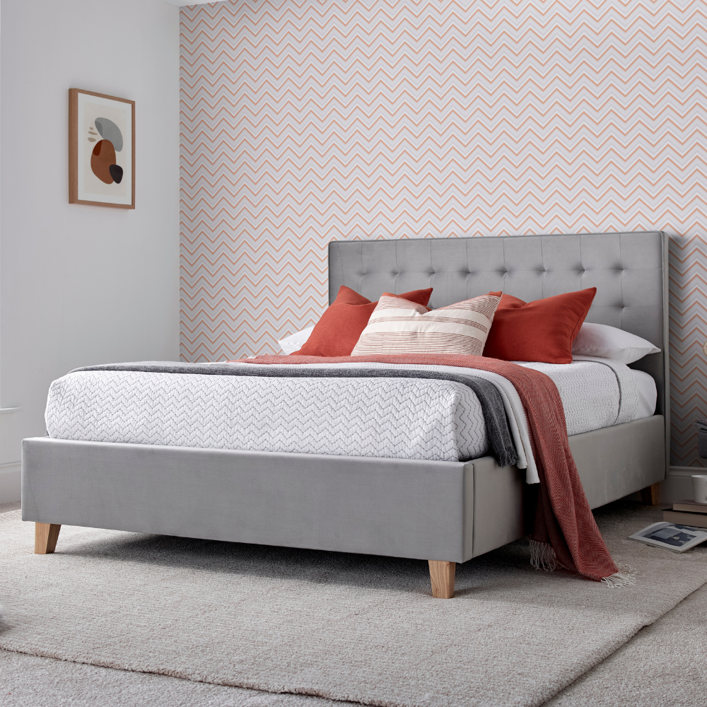 Grey upholstered bed frame in a room with pink patterned wallpaper.