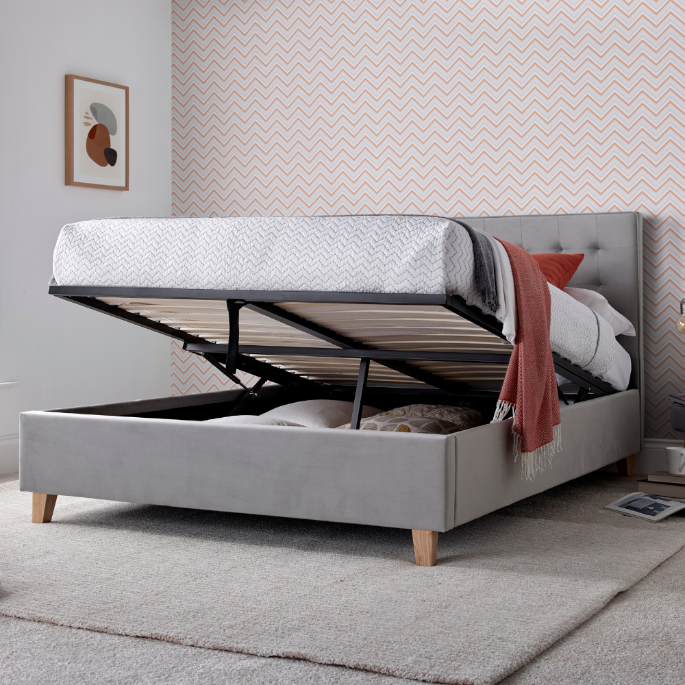 Grey upholstered ottoman bed frame opened up, in a bedroom with pink wallpaper.