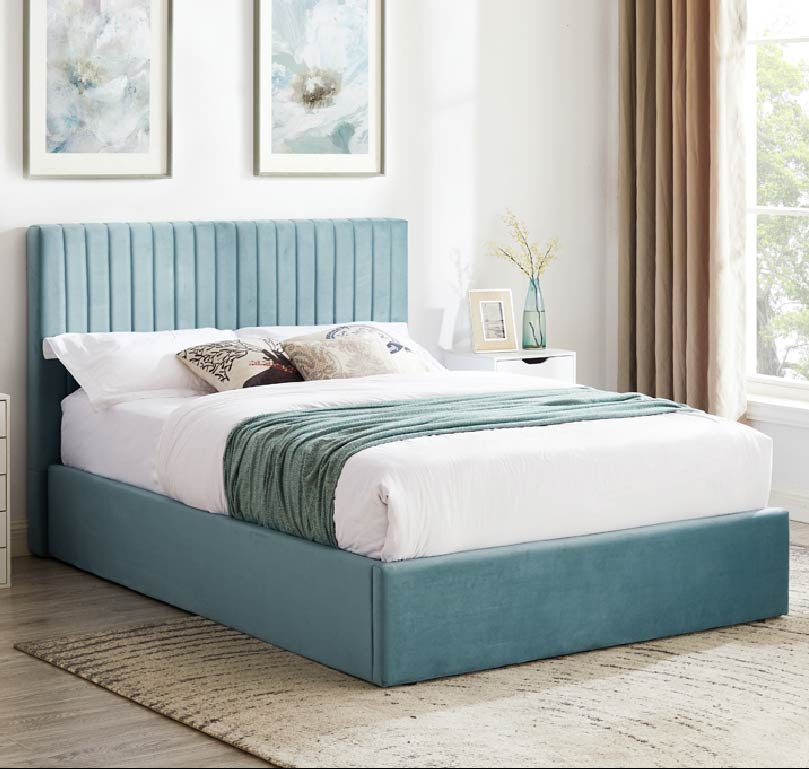 A blue velvet ottoman bed frame with a high, tufted headboard and storage space underneath, placed in a neatly arranged bedroom with decorative pillows on the bed.