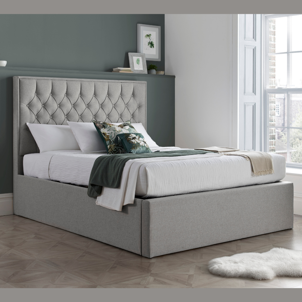 Grey upholstered bed with tufted headboard in a room with a window and framed pictures on the wall.