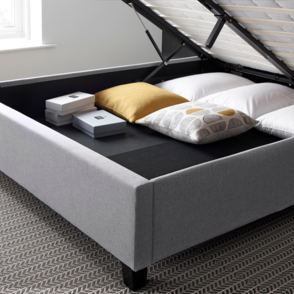 Grey upholstered bed with storage compartment containing pillows and boxes on a carpeted floor.