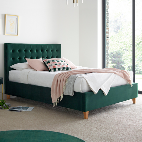 Green upholstered bed with white bedding and a pink blanket in a bright room.